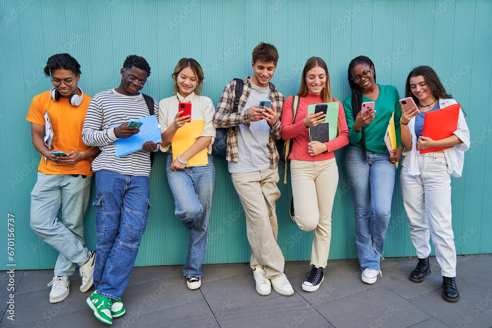 Fototapeta premium Cheerful multiracial group of young friends gathering to use phones while standing against a blue wall. Teenager students addicted to technology and social media looking at their own smartphones.