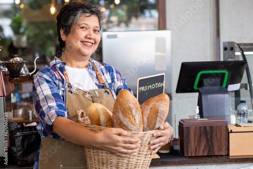 Retired businessman receiving a pension with gray hair Standing smiling, holding a basket of baked goods promotional sign on it. In front of the cafe's counter and bakery booth is a small business.