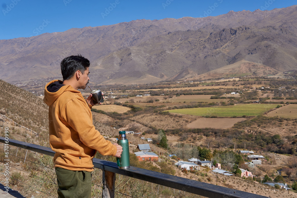 Naklejka premium latin man alone dressed in a mustard-colored sweatshirt, drinking mate looking at the landscape from a balcony in tafi del Valle, Tucuman, Argentina. Relaxed vacation