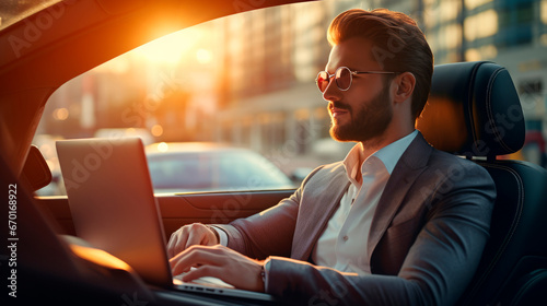 Handsome businessman in sunglasses and stylish costume, working with laptop in car flooded with sunlight, while going on business trip