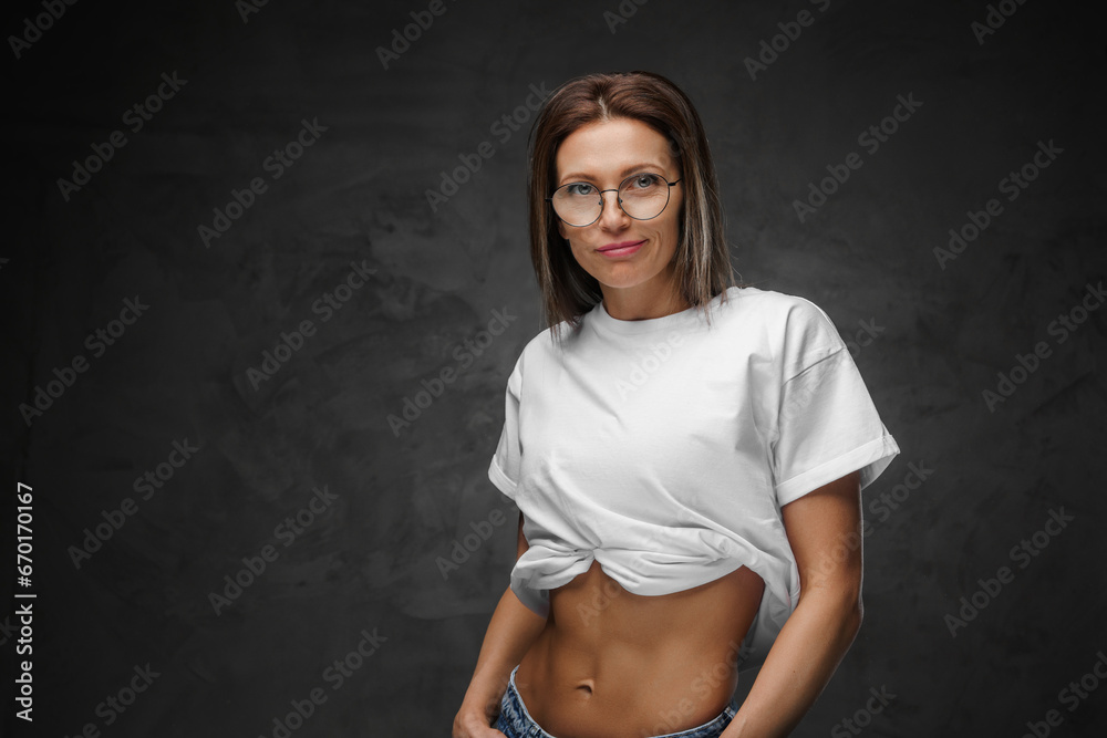 Obraz premium Alluring woman in glasses showcases her sculpted abs in a white crop-top against a dark backdrop