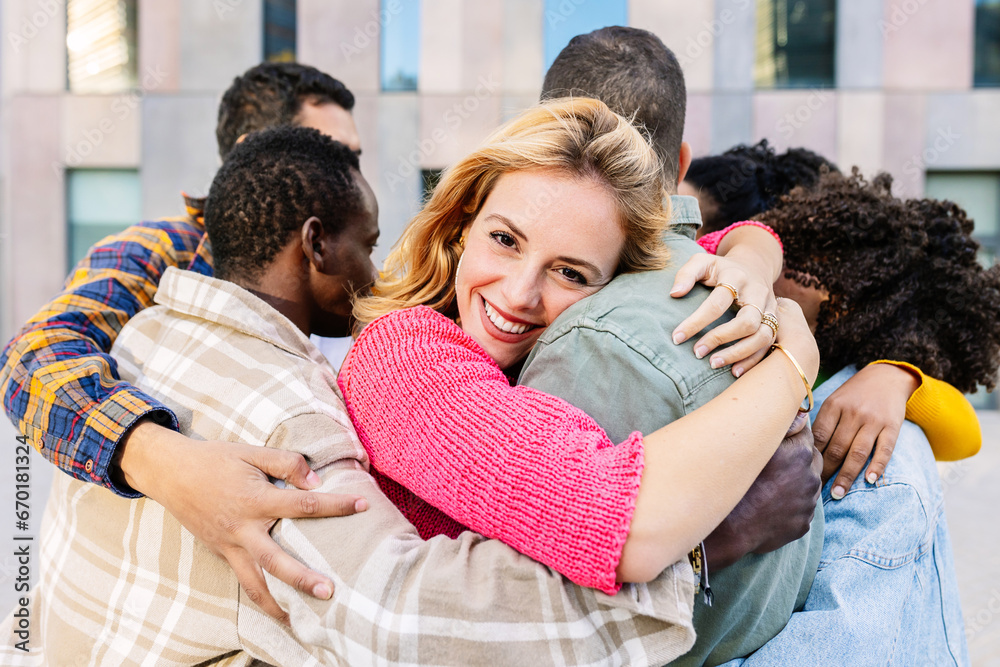 Young group of friends embracing together at city street. Beautiful woman smile at camera enjoying multiple hug with diverse friends. Love and friendship concept.