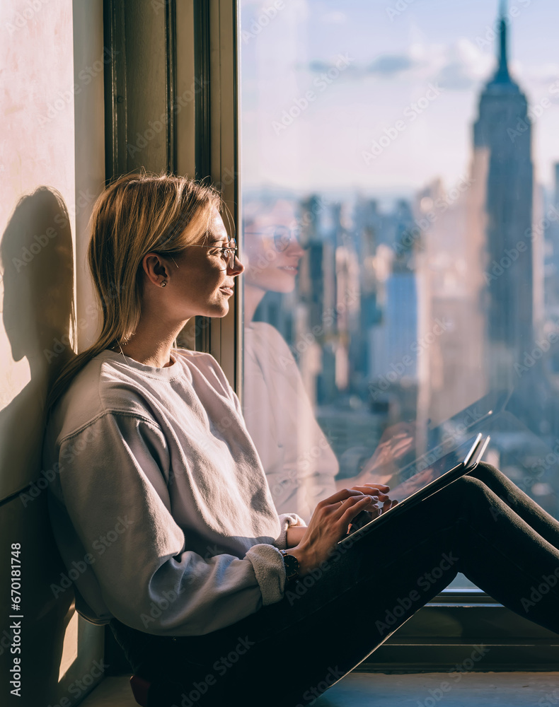 Fototapeta premium Young pensive female tourist inspired by scenery skyline of New York sitting on window