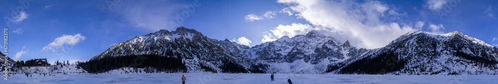 A panoramic view capturing the majestic snow-covered mountains under a clear blue sky. The frozen lake showcases visitors taking in the beauty, with a distant chalet providing a warm refuge.