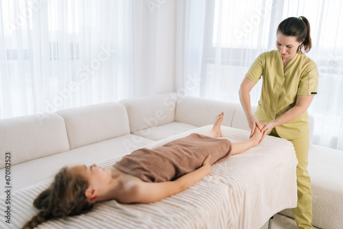 Wide shot of friendly female masseuse doing foot and legs massage to cute ten-year-old girl lying on massage table in spa salon. Little child having therapeutic, preventive massage session © dikushin