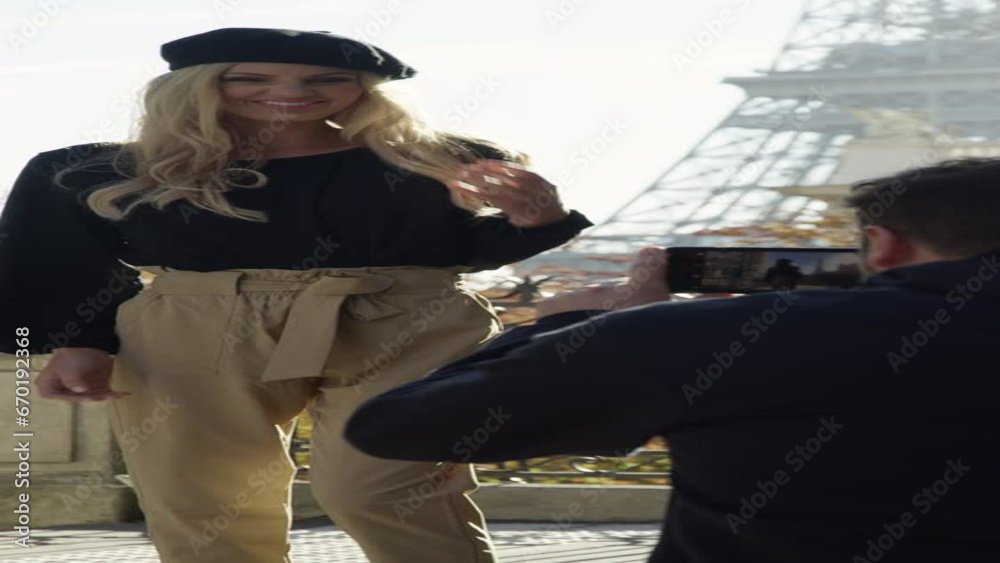 Crouching man with cell phone photographing woman posing near Eiffel Tower  - vertical video / Paris, Ile de France, France