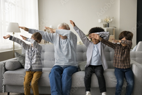 Older great-grandfather and three great-grandsons sit on couch in modern living room make dab popular youth culture gesture, multigenerational family perform triumph dance have fun together at home