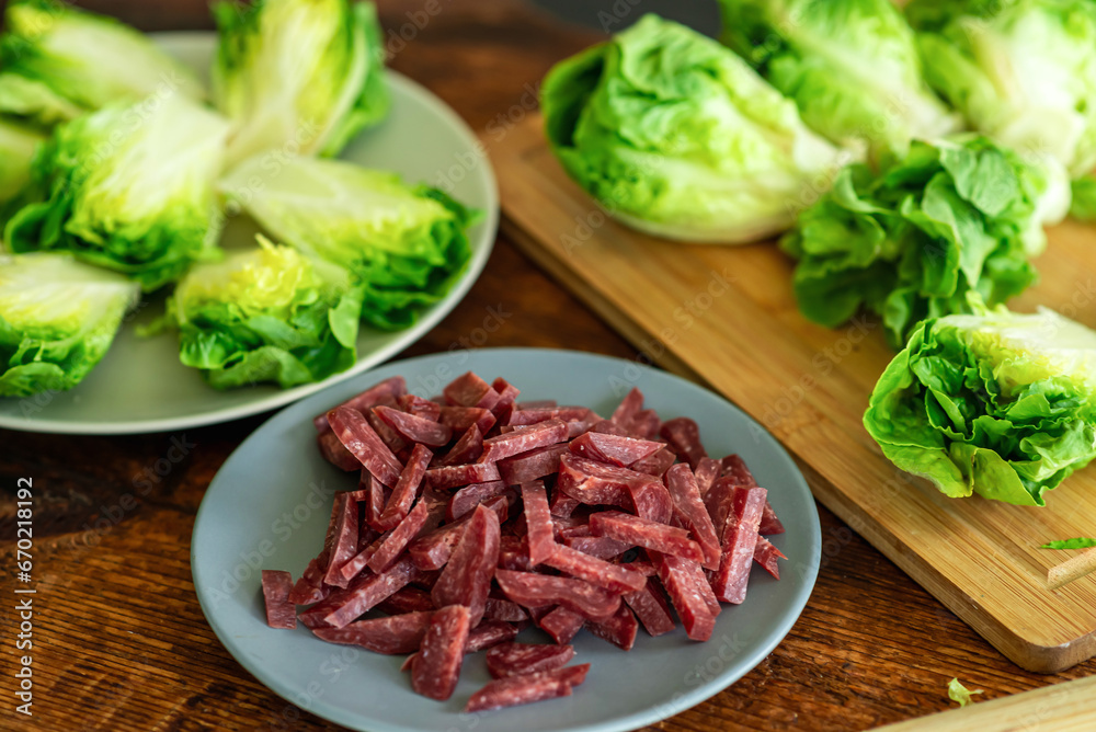 Fresh green lettuce cut into halves on a wooden cutting board. View from above. Chopped sausage. Recipe for stewed salad with meat. Fresh green lettuce cut into halves on a wooden cutting board. View from above. Chopped sausage. Recipe for stewed salad with meat.