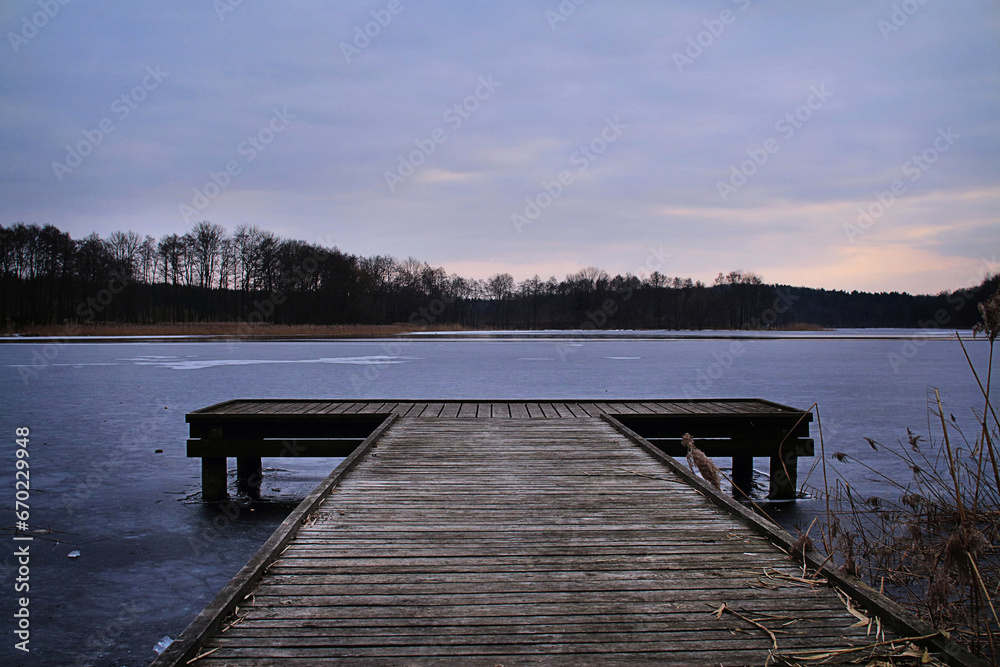 Drewniany molo na zamarzniętym jeziorze o zachodzie słońca. Zimowy wieczór. - obrazy, fototapety, plakaty 