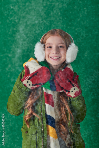 cheerful girl in ear muffs, striped scarf and winter attire standing under falling snow on turquoise