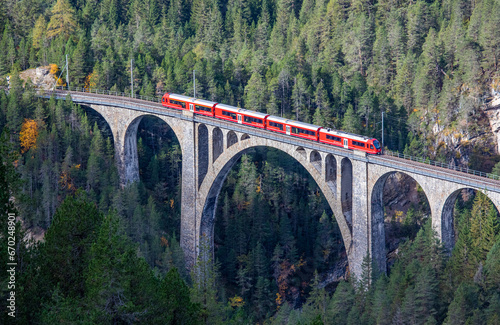 A red passenger train is crossing the famous Wiesener viaduct on the train line Davos - Filisur, the highest viaduct in swiss alps