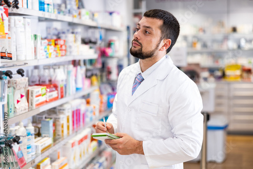 Friendly man pharmacist standing at drugstore with notepad and pen in hands