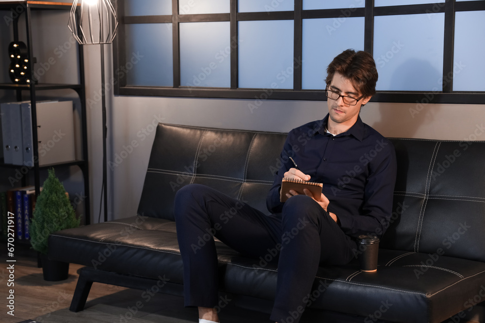 Young businessman sitting on sofa at office and writing in notebook