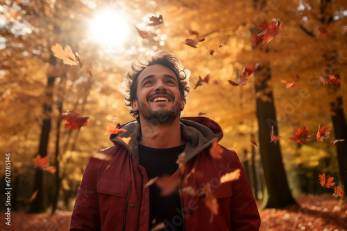 A young latin man is playing with fallen leaves happily with an autumn coat in a forest during sunset in autumn with a vibrant coloration
