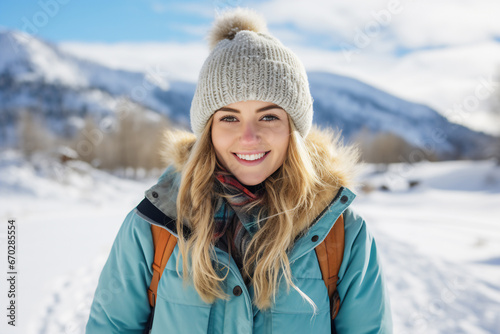 A young caucasian woman is posing in front of the camera happily with a winter coat and a winter hat in a in snow covered country landscape during day in winter on a bright day