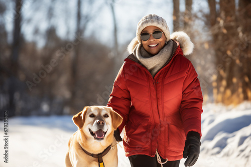 A senior african american woman is walking happily with the dog with a winter coat and winter hat in a in snow covered forest during day in winter on a bright sunny day