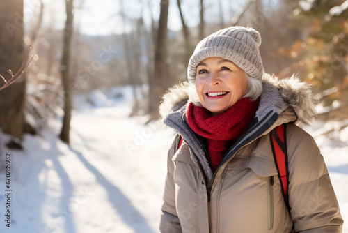A senior caucasian woman is walking happily with a winter coat and a winter hat in a in snow covered forest during day in winter on a bright day