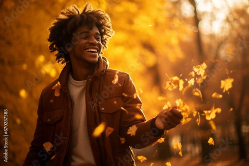 A young african american man is playing with fallen leaves happily with an autumn coat in a forest during sunset in autumn with a vibrant coloration