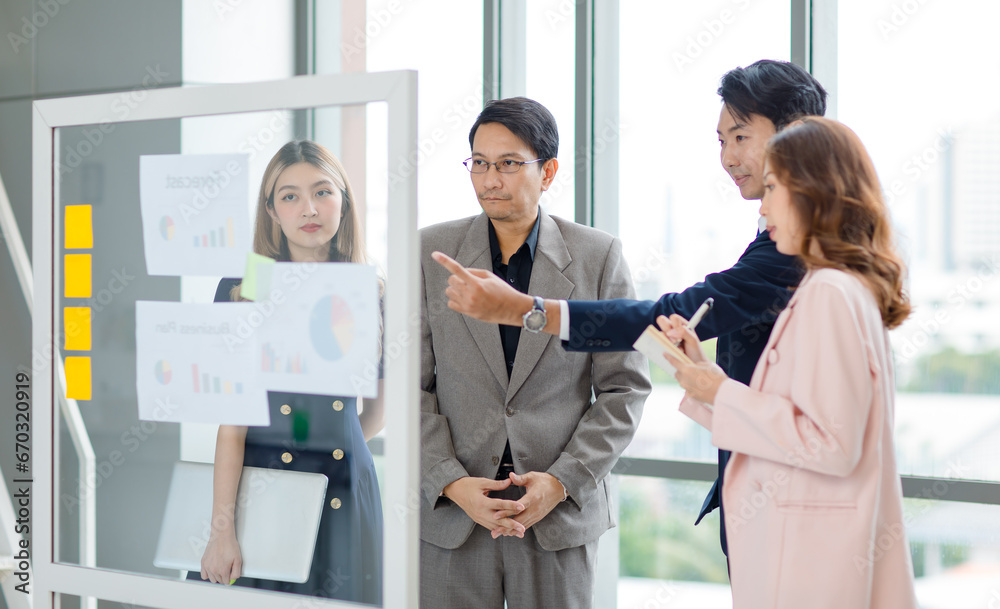 Obraz premium Asian professional successful young female businesswoman presenter lecturer in formal business suit outfit standing pointing color sticky note on glass board discussing with male colleague