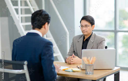 Asian professional successful male businessman interviewer recruiter in formal business suit outfit sitting interviewing unrecognizable applicant at working desk in company office recruiting room