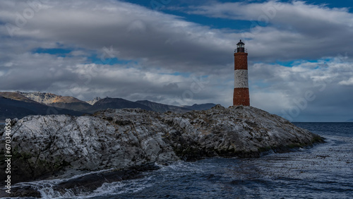 The famous southernmost old lighthouse at the end of the world. The red-and-white striped tower stands on a small rocky island in the Beagle Channel. The waves are beating against the cliff. 