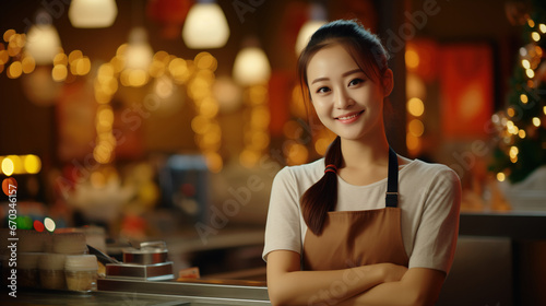 Young female barista smiling broadly  employee of a small local coffee bar  standing behind the counter with Christmas decorations and lights in the background. 