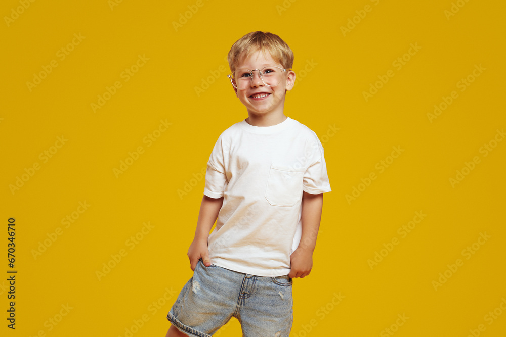 Little student boy in stylish white tshirt and eyeglasses smiling and looking at camera. Cute child laughing and standing isolated over yellow background - obrazy, fototapety, plakaty 