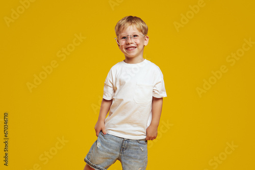 Little student boy in stylish white tshirt and eyeglasses smiling and looking at camera. Cute child laughing and standing isolated over yellow background photo