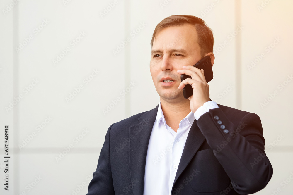 Confident adult businessman dressed in suit stands outdoors talking on phone. Concerned mature man in formal suit talks with interest on smartphone, sunlight Confident adult businessman dressed in suit stands outdoors talking on phone. Concerned mature man in formal suit talks with interest on smartphone, sunlight