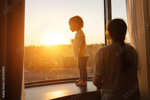 Happy parents with child stand together in front of window in modern flat at sunset. Successful family looks outside on sunrise from flat © lenblr