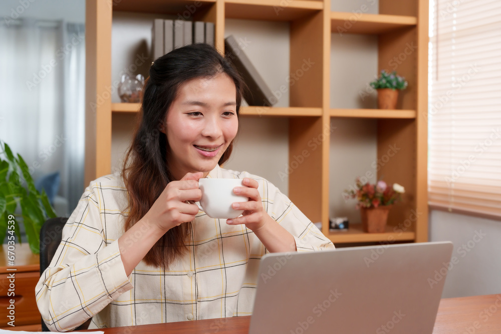 Close-up Asian woman hand holding hot coffee mug, while working in home office on laptop computer, placed in front desk, another hand is typing, working on laptop computer, happy smiling lips. Close-up Asian woman hand holding hot coffee mug, while working in home office on laptop computer, placed in front desk, another hand is typing, working on laptop computer, happy smiling lips.