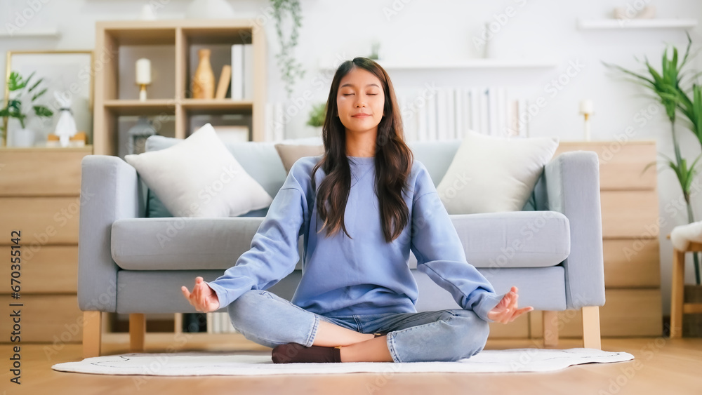 Happy young Asian woman practicing yoga and meditation at home sitting on floor in living room in lotus position and relaxing with closed eyes. Mindful meditation and wellbeing concept Happy young Asian woman practicing yoga and meditation at home sitting on floor in living room in lotus position and relaxing with closed eyes. Mindful meditation and wellbeing concept