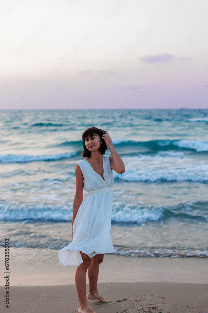 Happy woman in a white dress enjoys relaxing on the beach, walking along the sandy shore at sunset. Happy woman in a white dress enjoys relaxing on the beach, walking along the sandy shore at sunset.