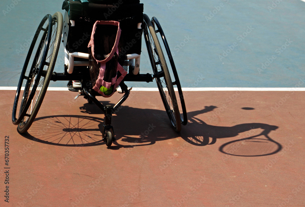 A person in a wheelchair plays tennis on the court. A disabled tennis player and his shadow hit the ball in an outdoor match.