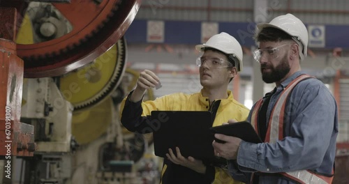 team of machinery factory engineers uses laptops to inspect machines that produce steel-related parts in a large factory. Working professionally, wearing hard hats to protect workplace safety.