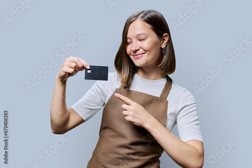 Young happy woman service worker in apron with credit card in hand, on gray background photo