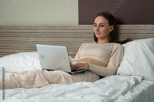 Young woman with long dark hair is lying on the bed and watching an interesting movie on a laptop at night at home.