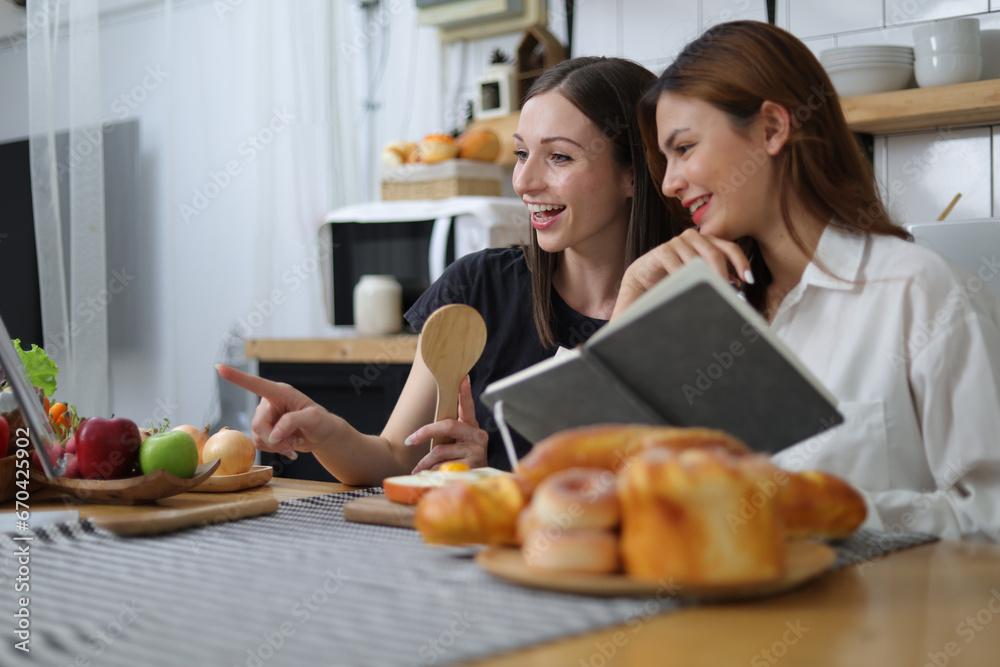 Two young women in the kitchen practicing cooking are looking at recipes on their tablets and notebooks. Two young women in the kitchen practicing cooking are looking at recipes on their tablets and notebooks.