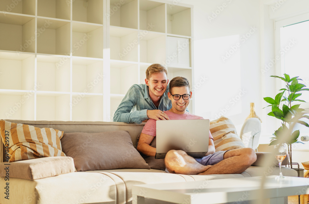 Young gay couple sitting in an apartment on a sofa using a laptop a and shopping online. Modern gay family Young gay couple sitting in an apartment on a sofa using a laptop a and shopping online. Modern gay family