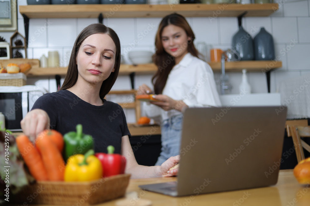 Two young women in the kitchen practicing cooking are looking at recipes on their laptops.