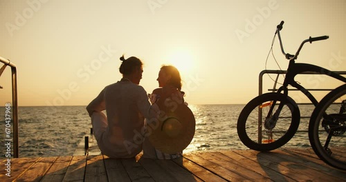 Zoom out guy and girl sitting on the pier on the beach opposite the sunrise and sea. Happy couple, a guy in light clothes and a girl with a Straw Hat, arrived on bicycles at Sunrise and are sitting photo