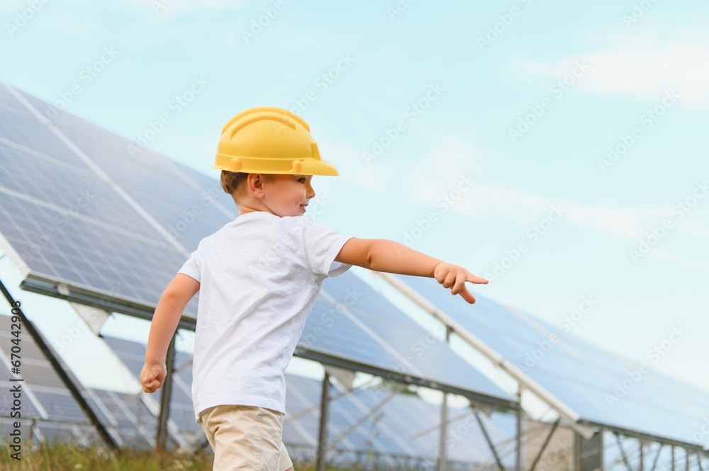 A happy little boy in a yellow helmet is standing on a solar panel farm.