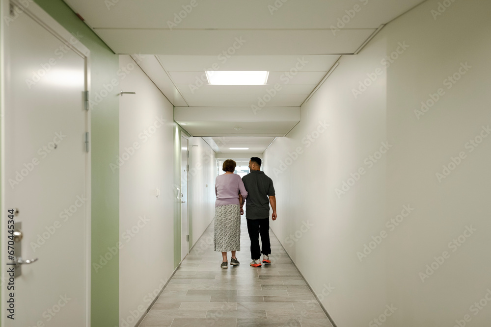 Full length rear view of senior woman walking arm in arm with male nurse at retirement home corridor Full length rear view of senior woman walking arm in arm with male nurse at retirement home corridor