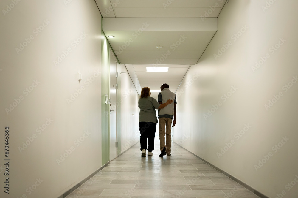 Full length rear view of female caregiver walking with arm around senior man in corridor at nursing home Full length rear view of female caregiver walking with arm around senior man in corridor at nursing home