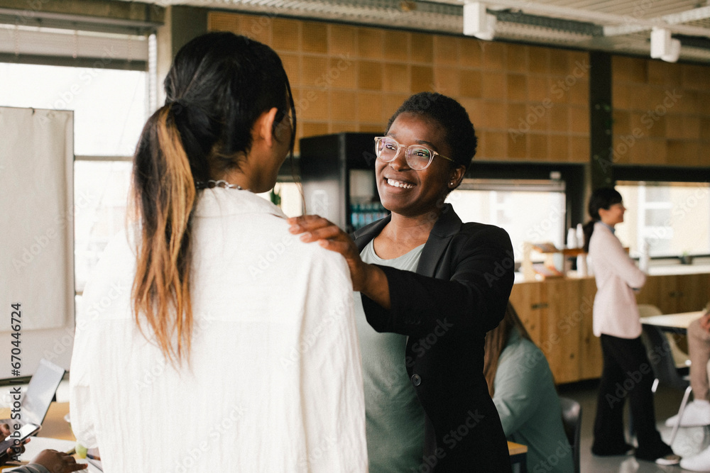 Happy businesswoman with hand on non-binary colleague's shoulder at office Happy businesswoman with hand on non-binary colleague's shoulder at office