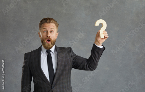 Businessman finds answer and business solution. Portrait of happy funny looking curious bearded man in suit with surprised face expression holding question mark standing isolated on grey background