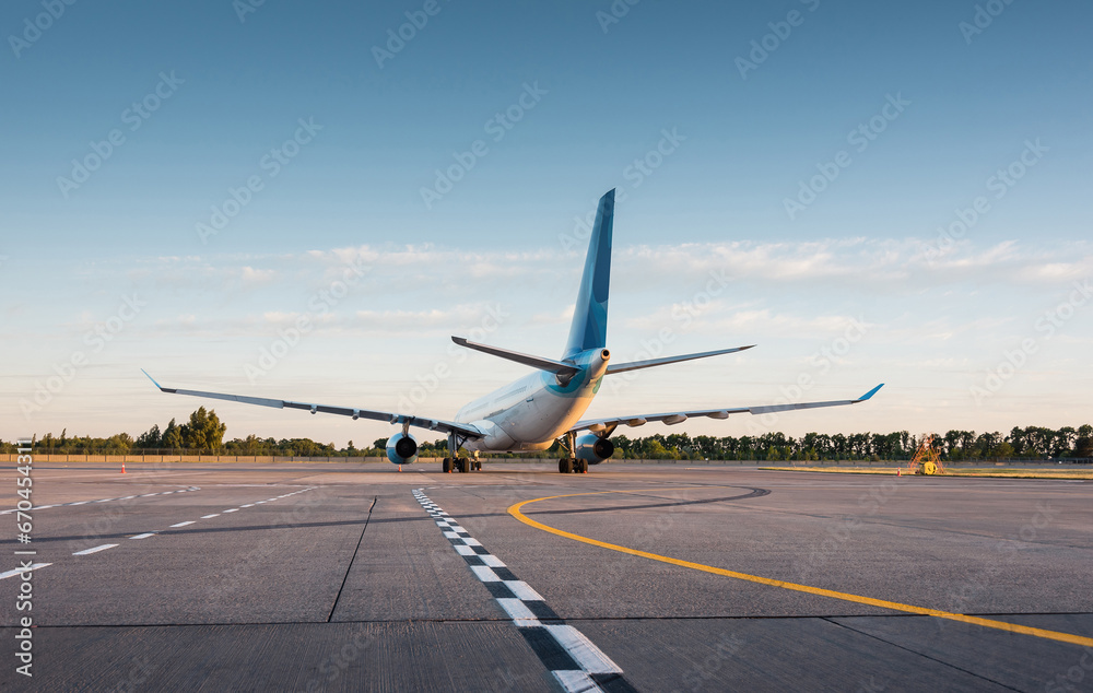 Obraz premium Rear view of white modern widebody passenger airplane on airport apron at sunset. Empty concrete apron with road markings and lines in the bottom third of the image.