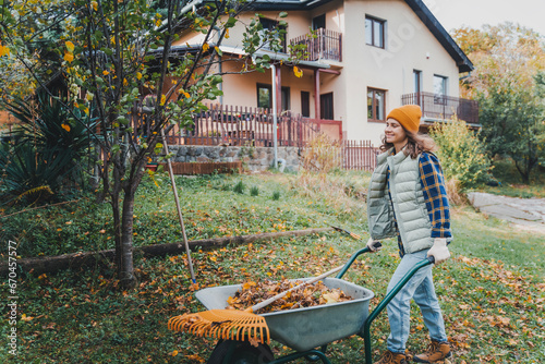 Young happy caucasian woman collecting autumn leaves in the backyard of a country house using a rake and cart