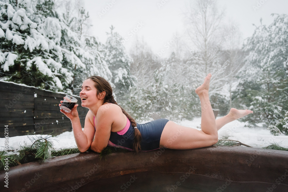 Woman drink cool drinks and relax in hot bath outdoors. Female enjoying thermal spa snowy forest. Winter holidays in mountains, hot water treatments concept. Young girl in open air while snowing.