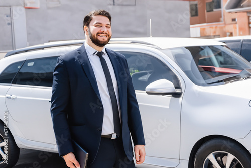 a man stands near the car. happy bearded man in a black business suit stands near his big white car in the city center